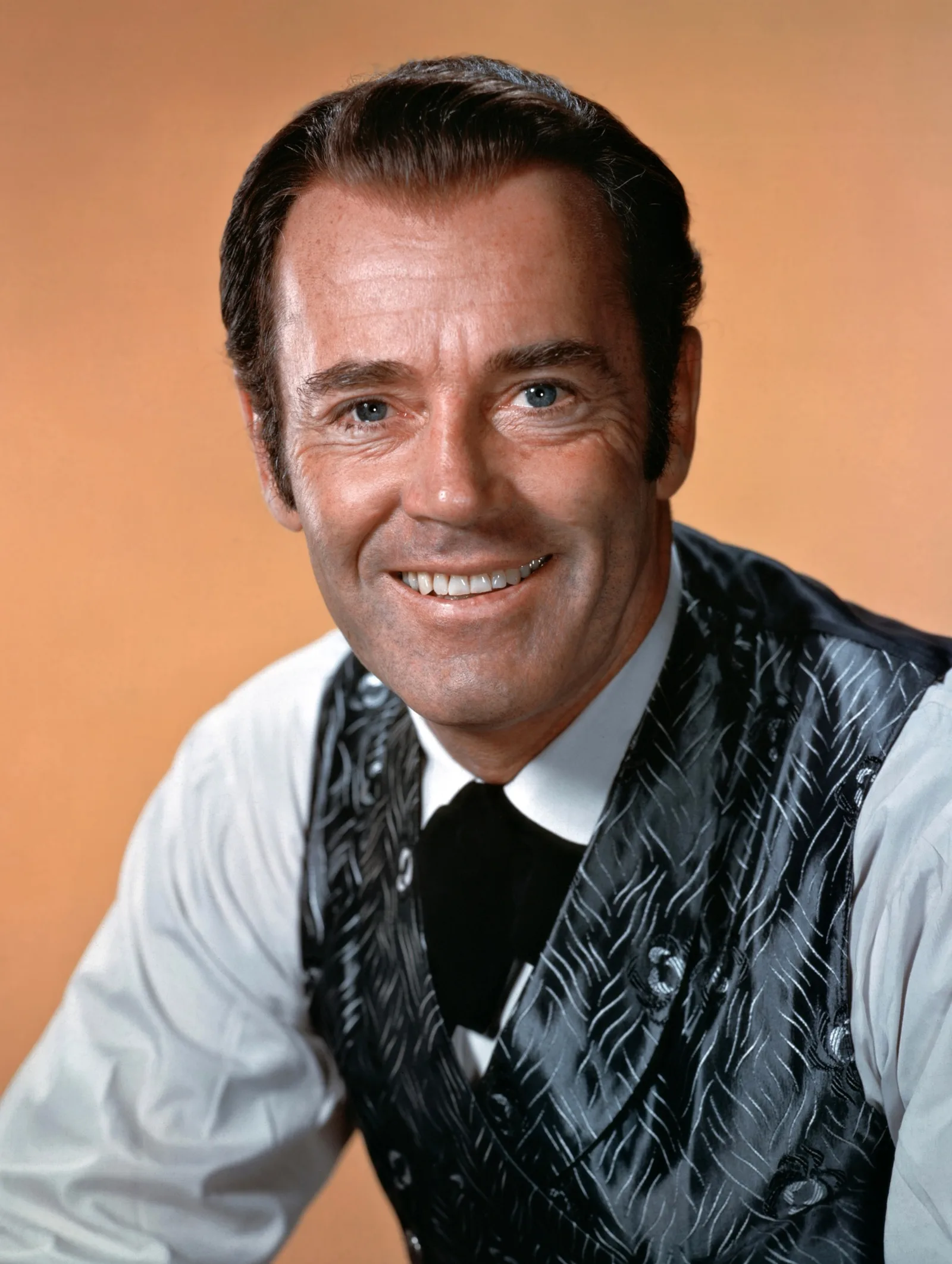 Colour studio publicity portrait of Henry Fonda smiling in a patterned grey waistcoat, white shirt and black cravat against a warm orange backdrop, for the 1959 film Warlock.