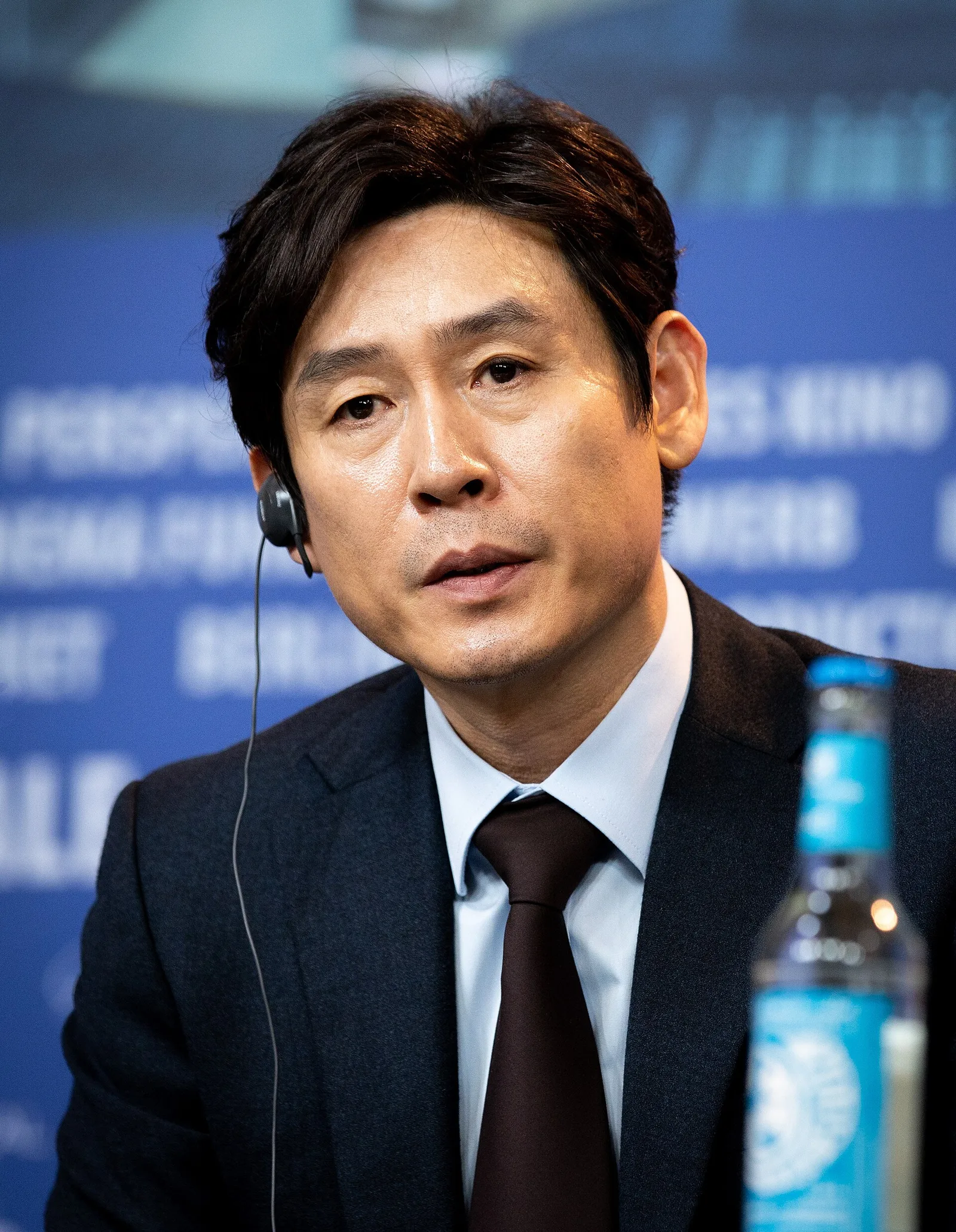 Actor Sol Kyung-gu in a navy suit and brown tie, wearing an interpreter's earpiece, speaking at a press conference at the 2019 Berlinale.