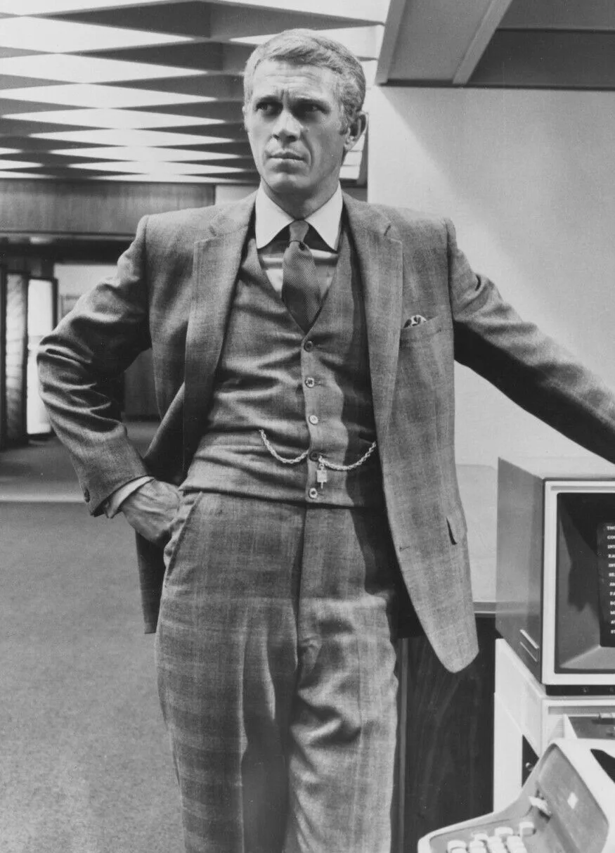 Black-and-white full-length still of Steve McQueen in a three-piece checked suit with a watch chain, one hand on his hip, standing in an office interior, 1968.