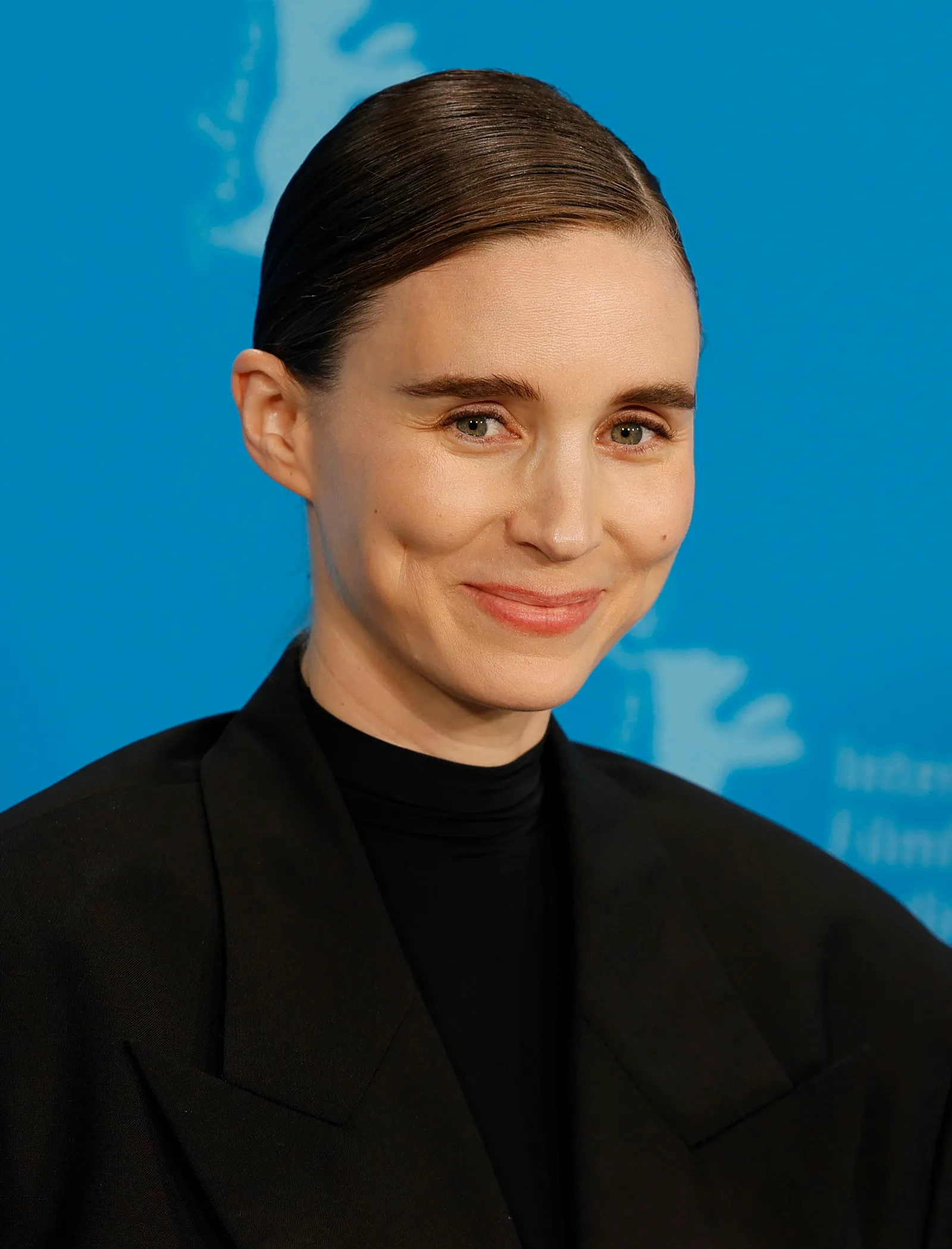 Rooney Mara smiling slightly, hair pulled back flat, in a black turtleneck and oversized black blazer against the blue Berlinale photocall backdrop, 2024.