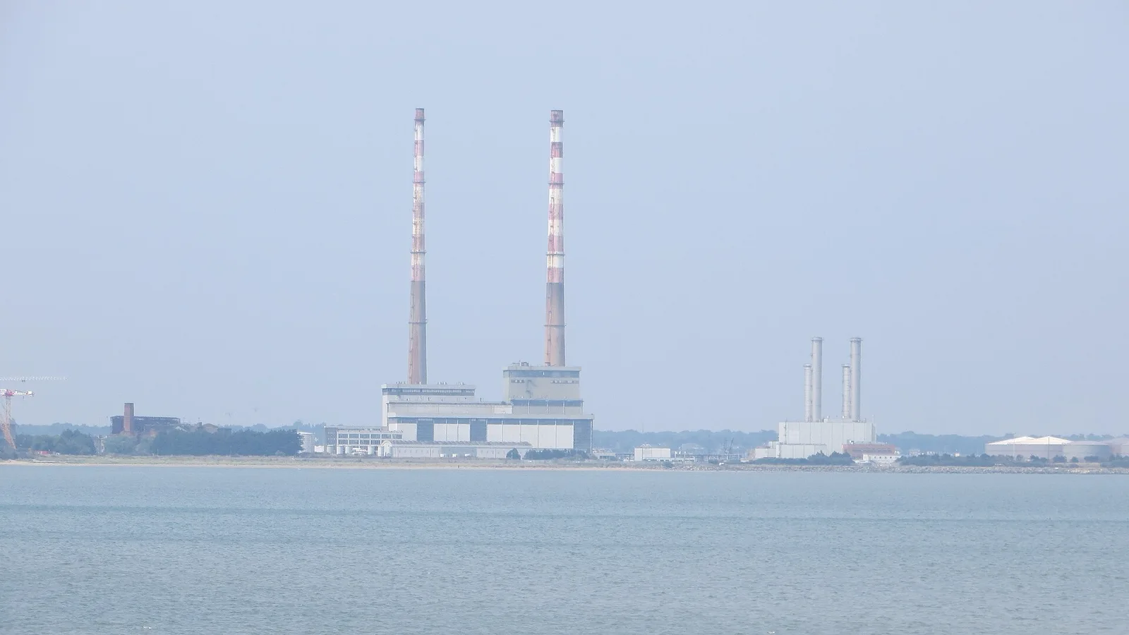 The two red-and-white banded Poolbeg chimneys rising over the low generating station on the Dublin Bay shoreline, seen across flat grey water from Booterstown in hazy light, July 2022.