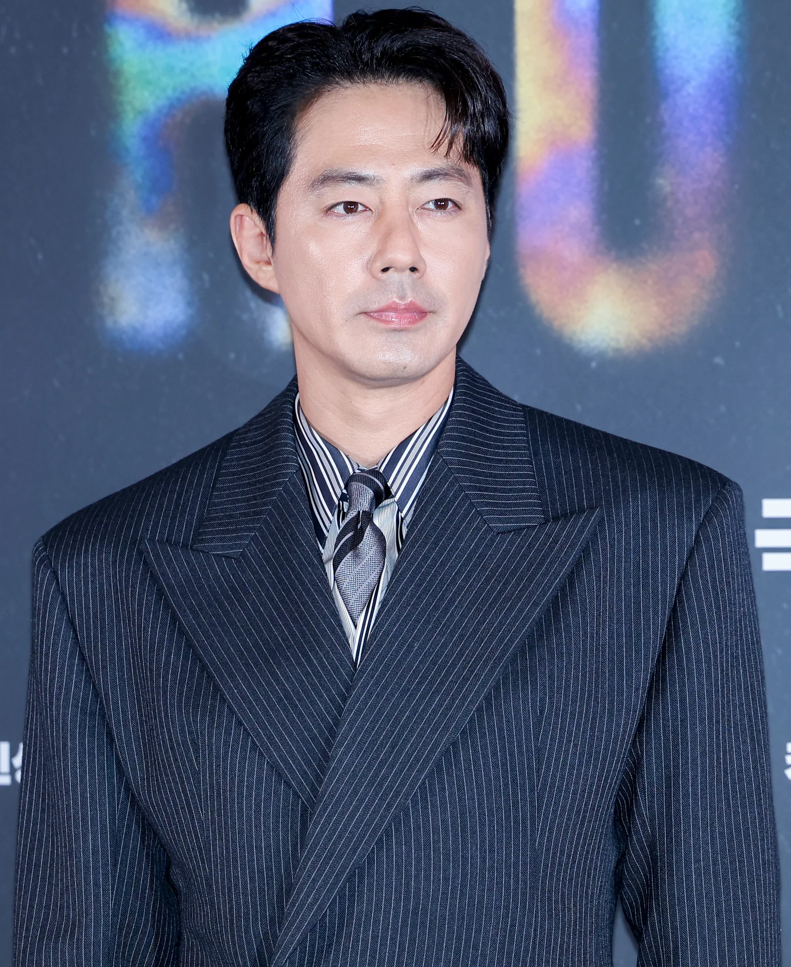 Actor Zo In-sung in a wide-shouldered navy pinstripe suit and grey tie, photographed against a dark event backdrop.