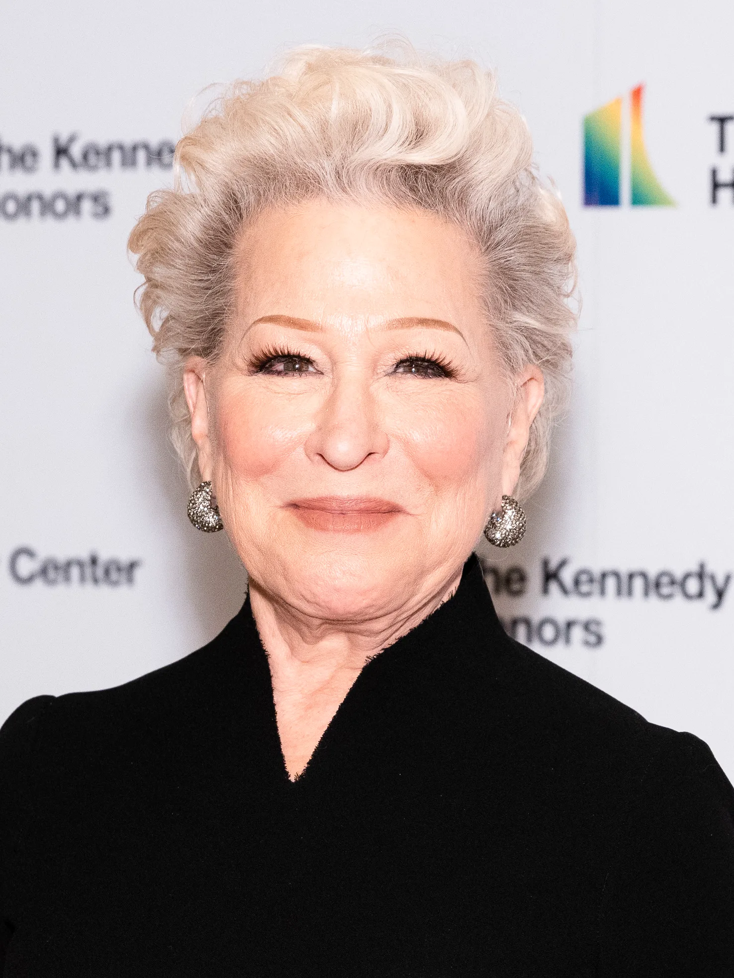 Bette Midler smiling in a high-necked black dress and silver drop earrings in front of a Kennedy Center Honors backdrop at the 2021 Medallion Ceremony.