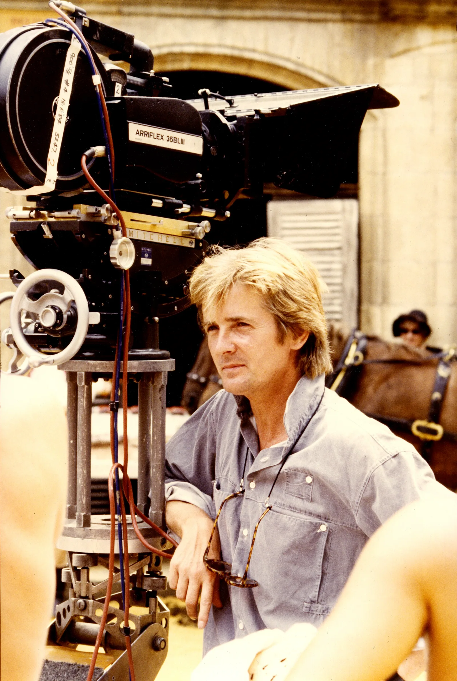 The director Franc Roddam standing beside an Arriflex film camera on set.