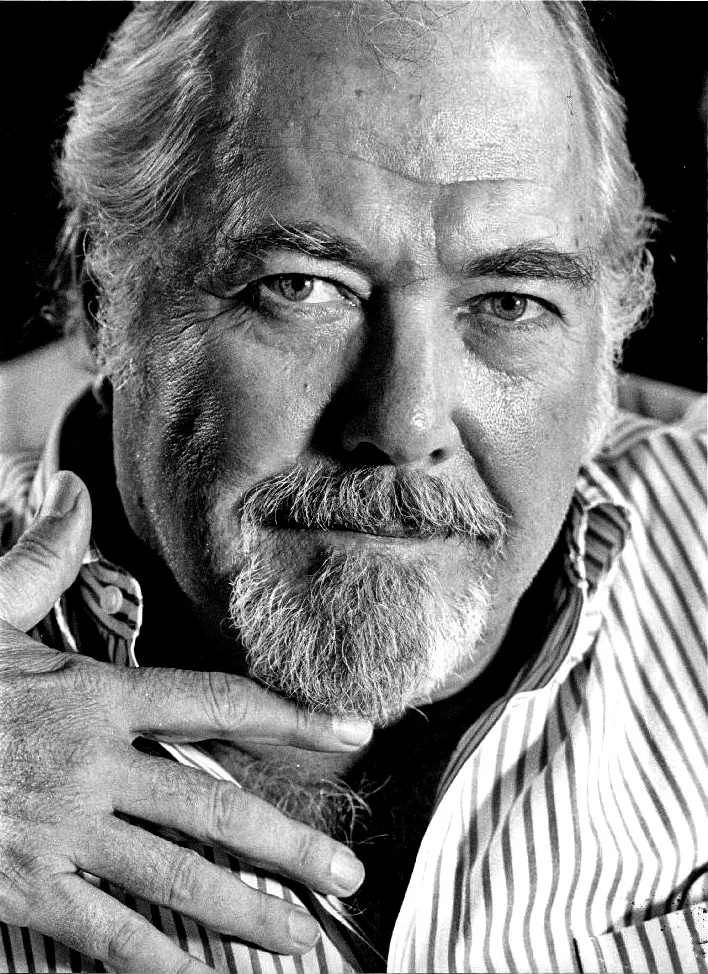 Close black-and-white publicity portrait of Robert Altman in 1983, white hair and full white beard, a striped shirt open at the collar, one hand raised against his cheek.