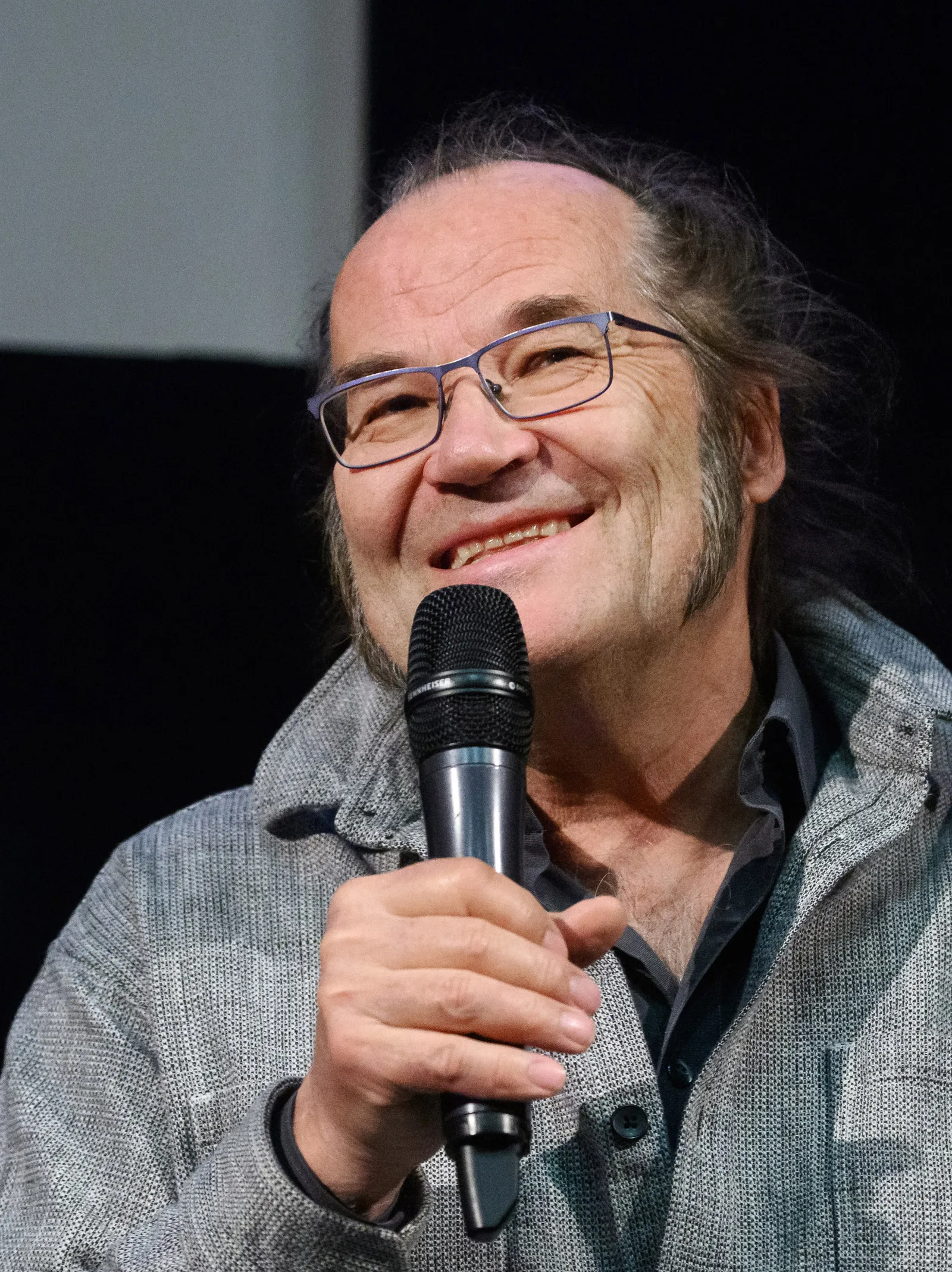 Cinematographer Peter Zeitlinger laughing while holding a handheld microphone, in glasses and a grey jacket, at a screening and talk at the Slovenian Cinematheque in Ljubljana, 2026.
