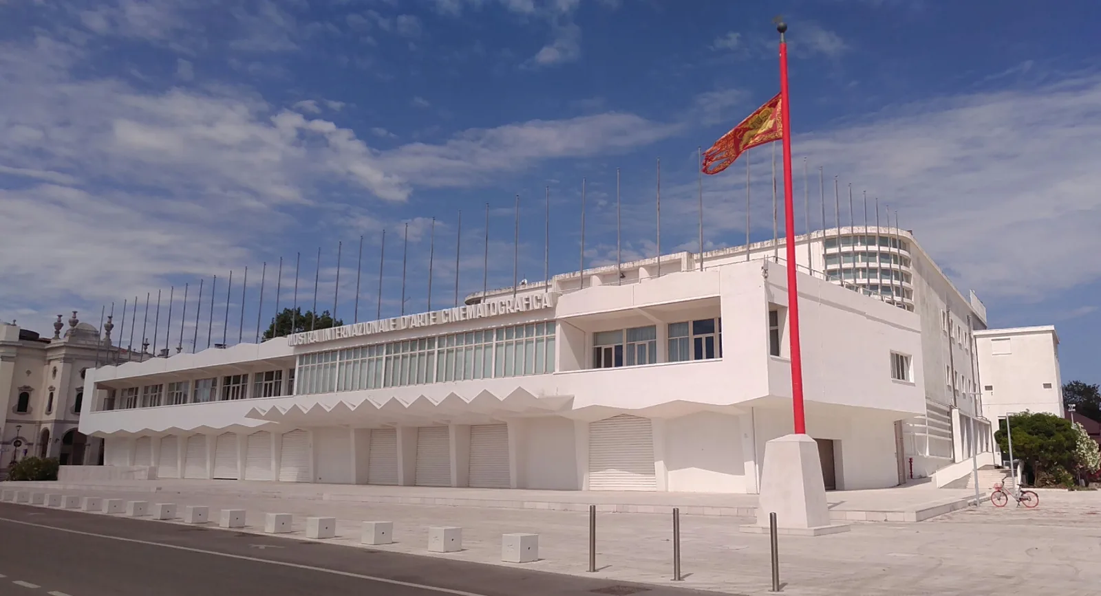 The white modernist Palazzo del Cinema on the Lido di Venezia under a blue sky, its row of bare flagpoles along the roofline and a tall red Venetian banner beside the entrance, 2021.
