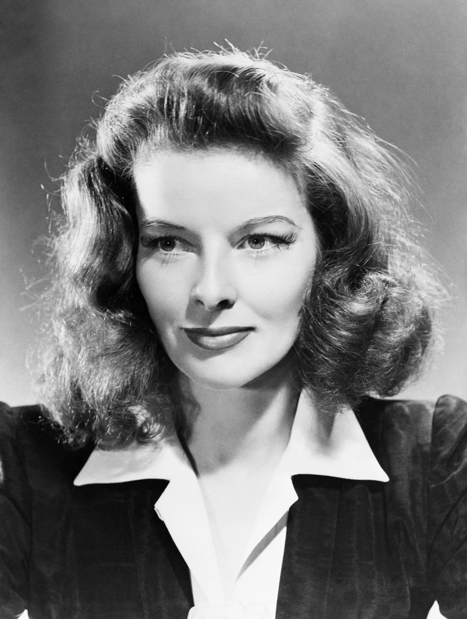 Black-and-white MGM studio publicity portrait of Katharine Hepburn, shoulder-length waved hair, in a dark jacket with a wide white collar, looking straight at the camera, 1941.